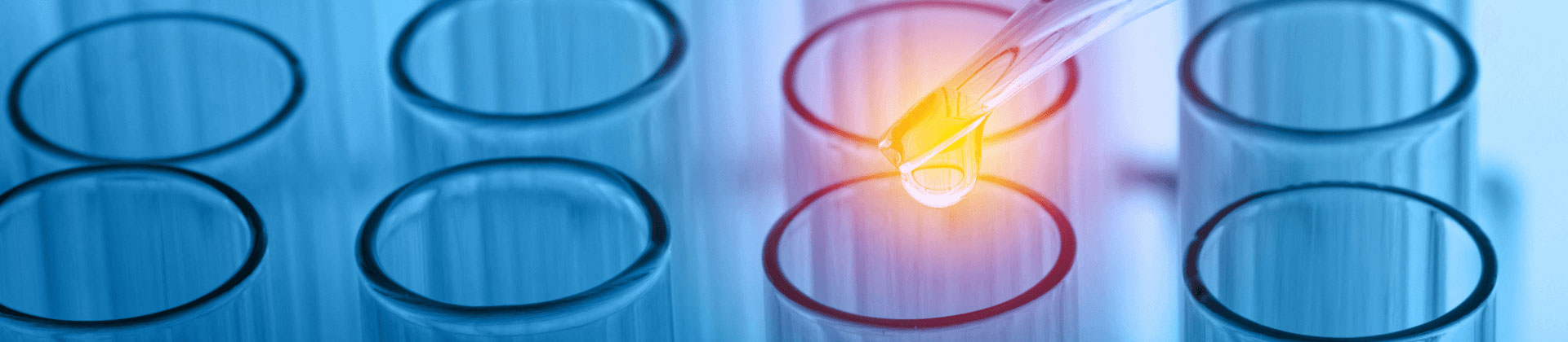 Close up of a glass pipette dropping gold colored liquid into the top of a set of glass chemistry vials.